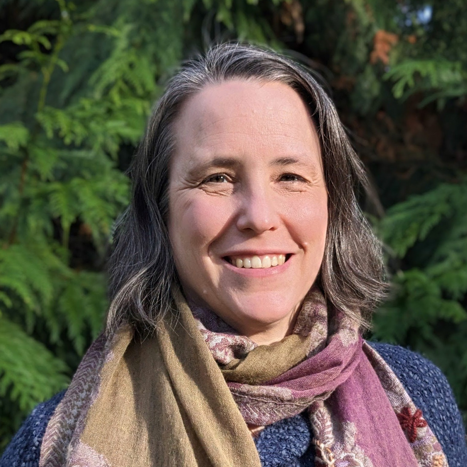 close up photo of a white female smiling in front of a cedar tree on a sunny day. She is wearing a comfy wool shawl and has straight short brown hair with grey highlights.